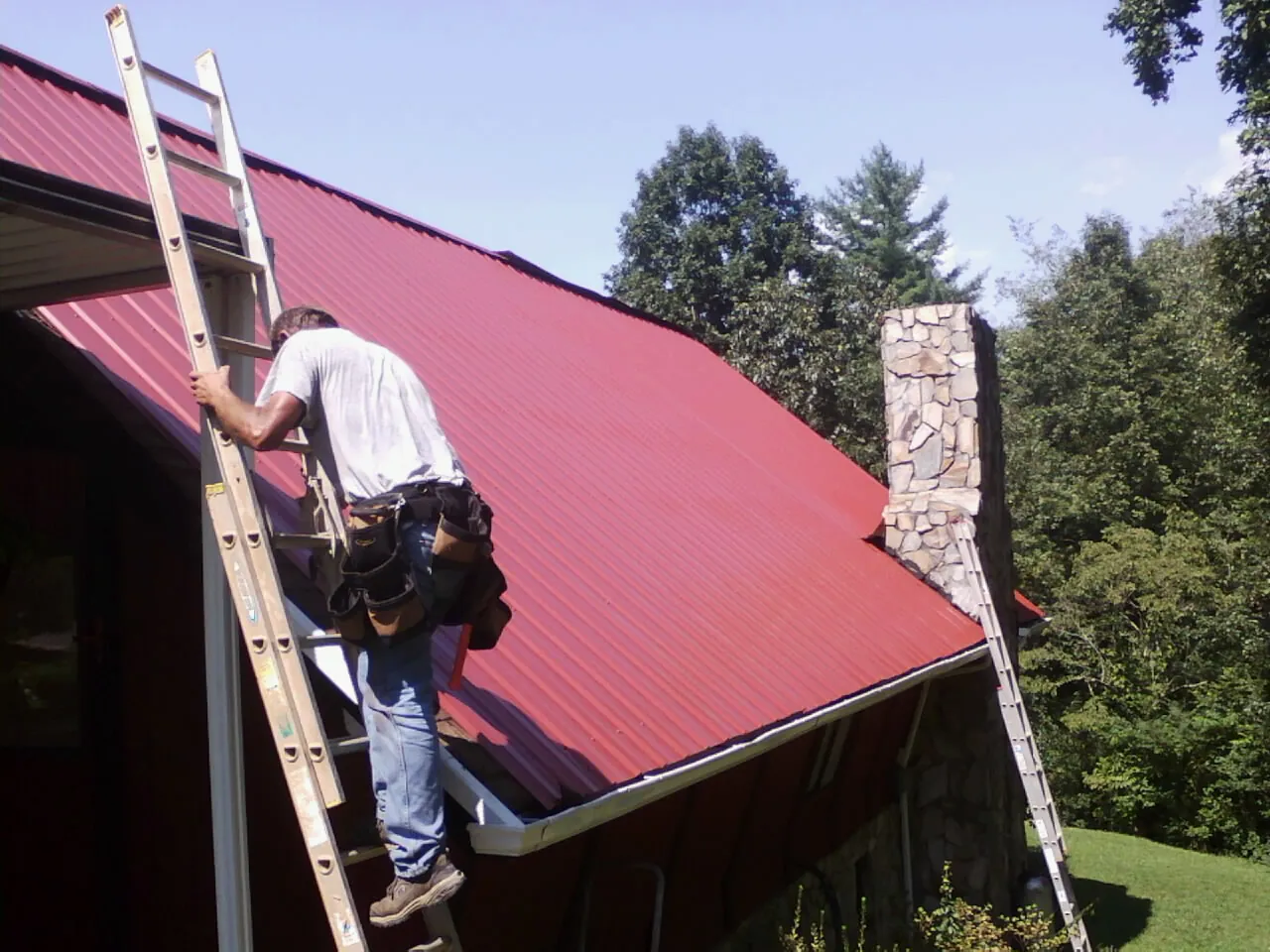 Roofer on a ladder beside a red metal roof and stone chimney — Soffit & Fascia Repair by Roofing Brothers in Chickamauga